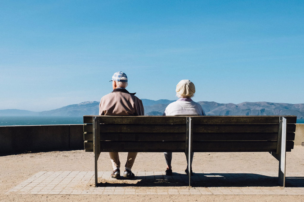 Elderly couple sitting on bench looking at mountain landscape.