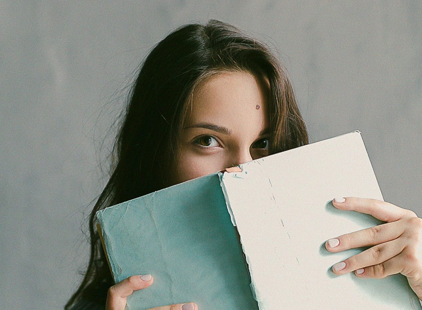 Shy woman hides half her face behind a pale blue book