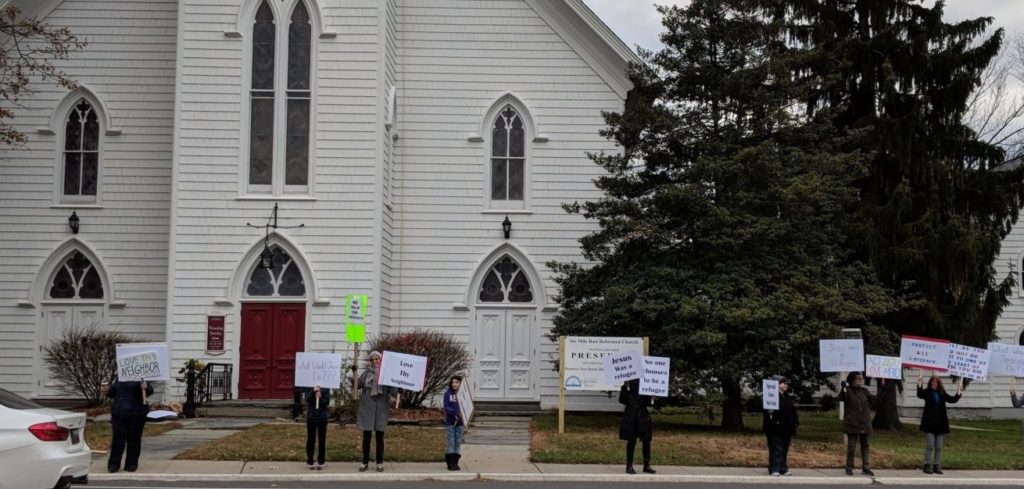 A small crowd of people stand on the sidewalk outside of a historic-looking church building, holding signs that express support of immigrants
