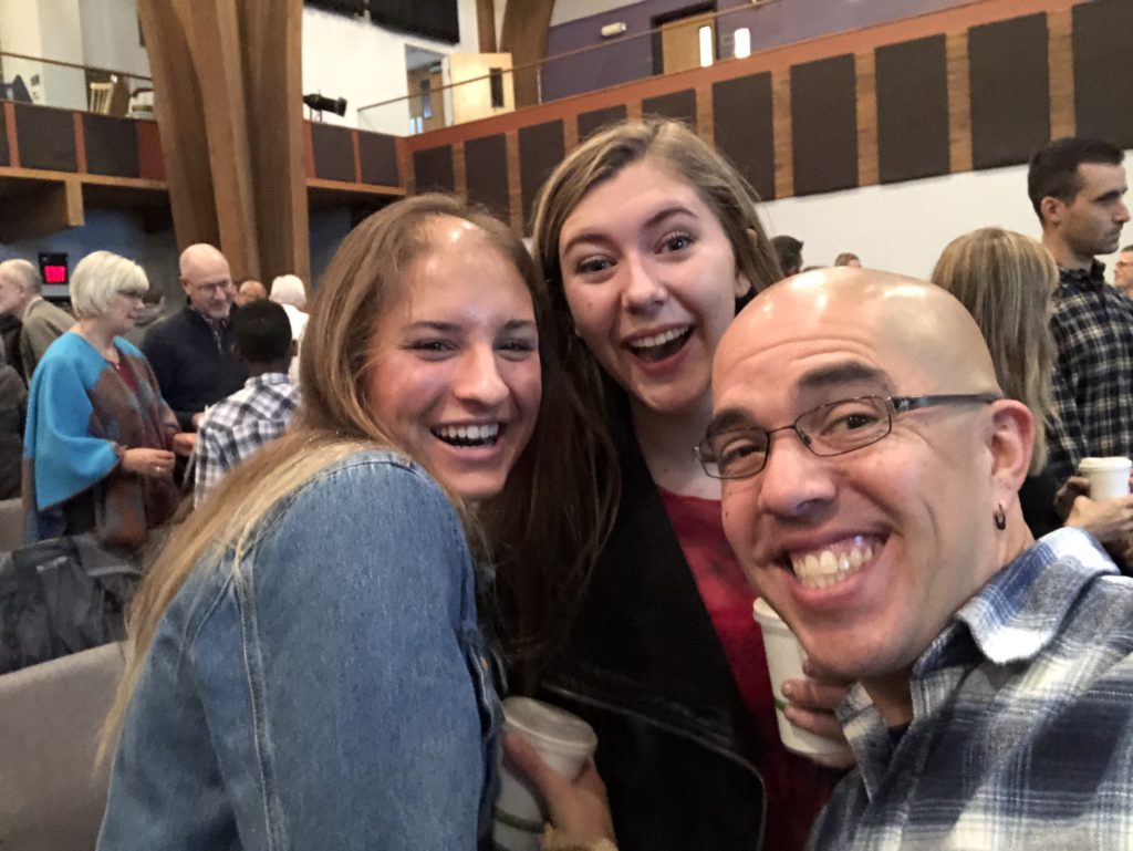 Two college women and a 40-something man with glasses and a shaved head pose for a selfie in a church sanctuary