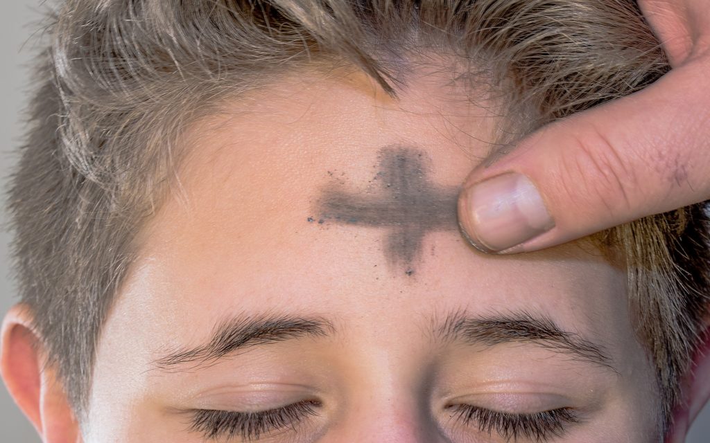 Un pulgar barre un trazo horizontal de ceniza en la frente de un joven, completando la señal de la cruz.