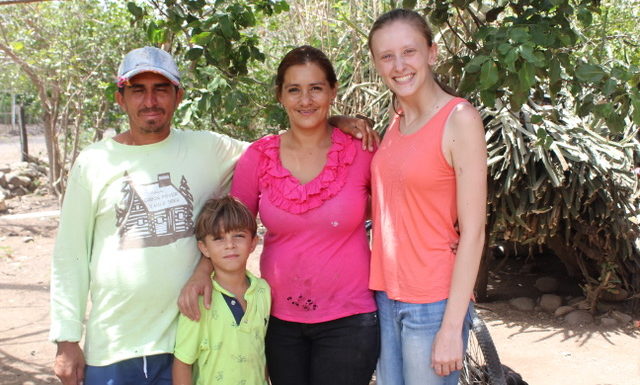 A Nicaraguan family and a young American woman, all wearing colorful clothing, stand under a tree on a sunny day