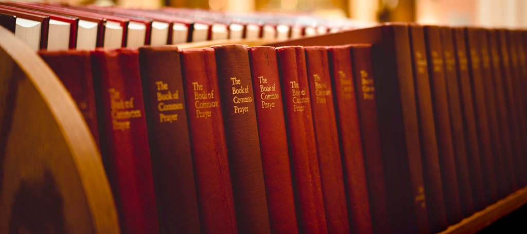 Los ejemplares de un libro de tapas rojas están colocados en fila en una estantería de madera. En la portada se lee: The Book of Common Prayer.