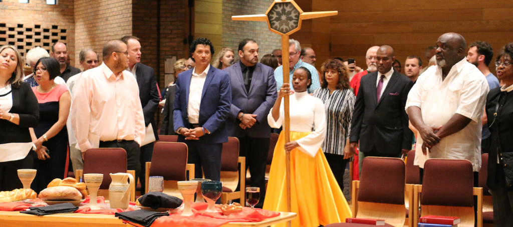 A confident young woman in flowing dancer’s attire carries a cross into a worship space as worshipers look on.