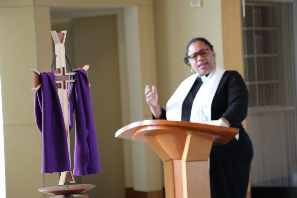 Black woman preaching at wooden podium with cross draped in purple in the foreground