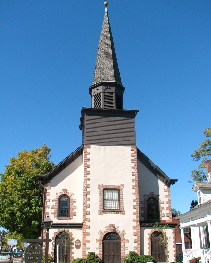 A tan church with wooden doors and a gray steeple stands tall against the clear blue sky.