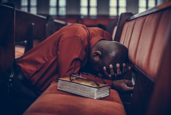 A man with his head in his hands kneels on a church pew next to a Bible.