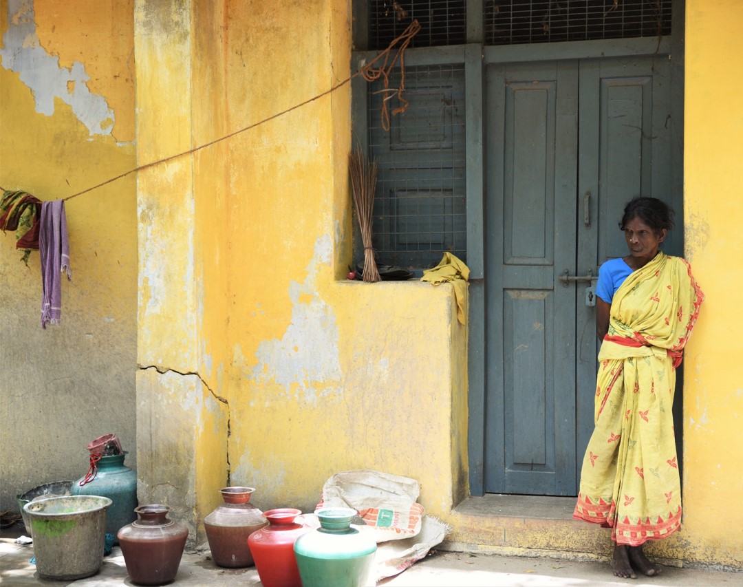 An Indian woman in a yellow sari leans against a cement wall, empty water jugs in front of her.