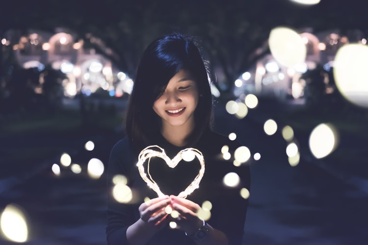 A young Asian woman holds a glowing heart-shaped string of lights with glimmers that extend across the dark background.