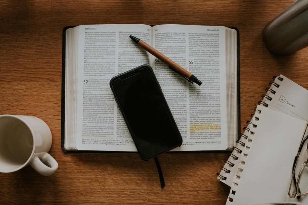 A phone and pen lie on top of an open Bible with highlighting. A journal and empty mug are beside.
