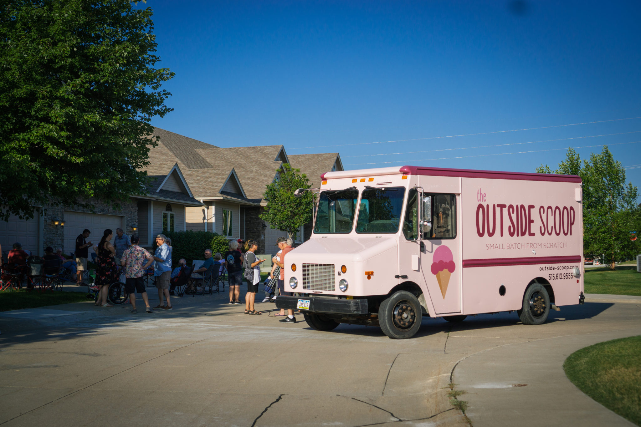 A light pink ice cream truck is parked on the street with a line of families being served.
