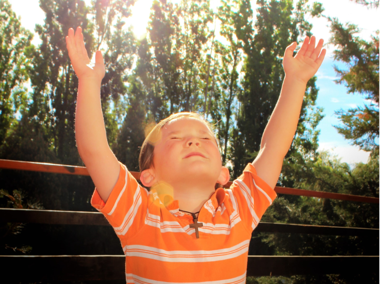 a young boy with an orange polo and a wooden cross necklace lifts his arms in prayer