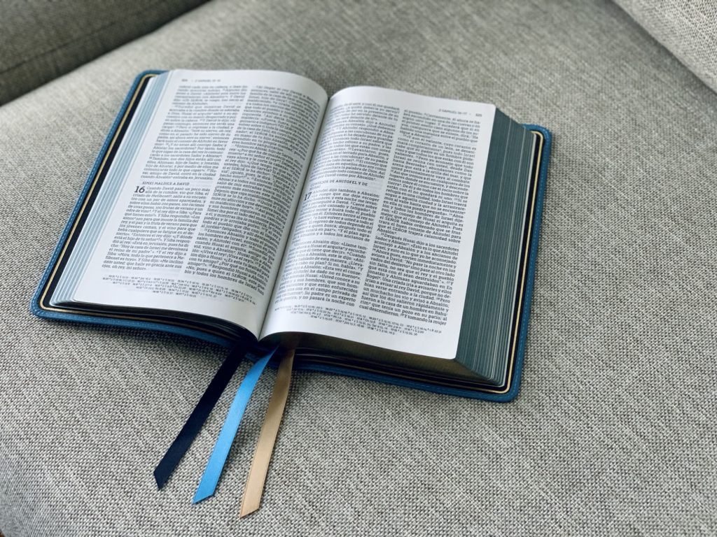 a blue leather bound Bible with multi colored ribbon bookmarks is open on a gray sofa