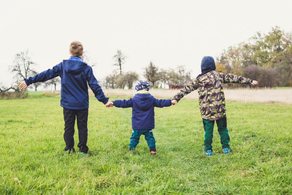 three kids in coats hold hands while standing on grass