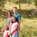 a man and woman stand with their two children on a grassy plain with a zebra in the background