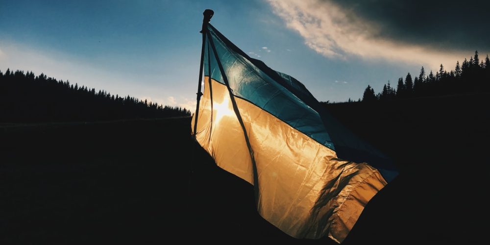the Ukrainian flag blows in the wind, backlit by the sun and dark hills