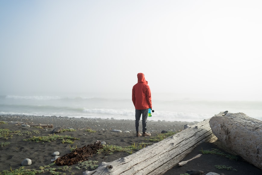 un hombre con abrigo rojo se encuentra en la orilla de la arena con barreras de troncos de árboles a su lado