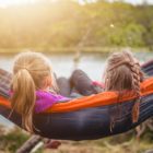 two girls relax in a nylon hammock overlooking lake and trees