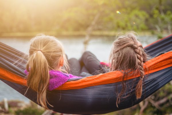 two girls relax in a nylon hammock overlooking lake and trees