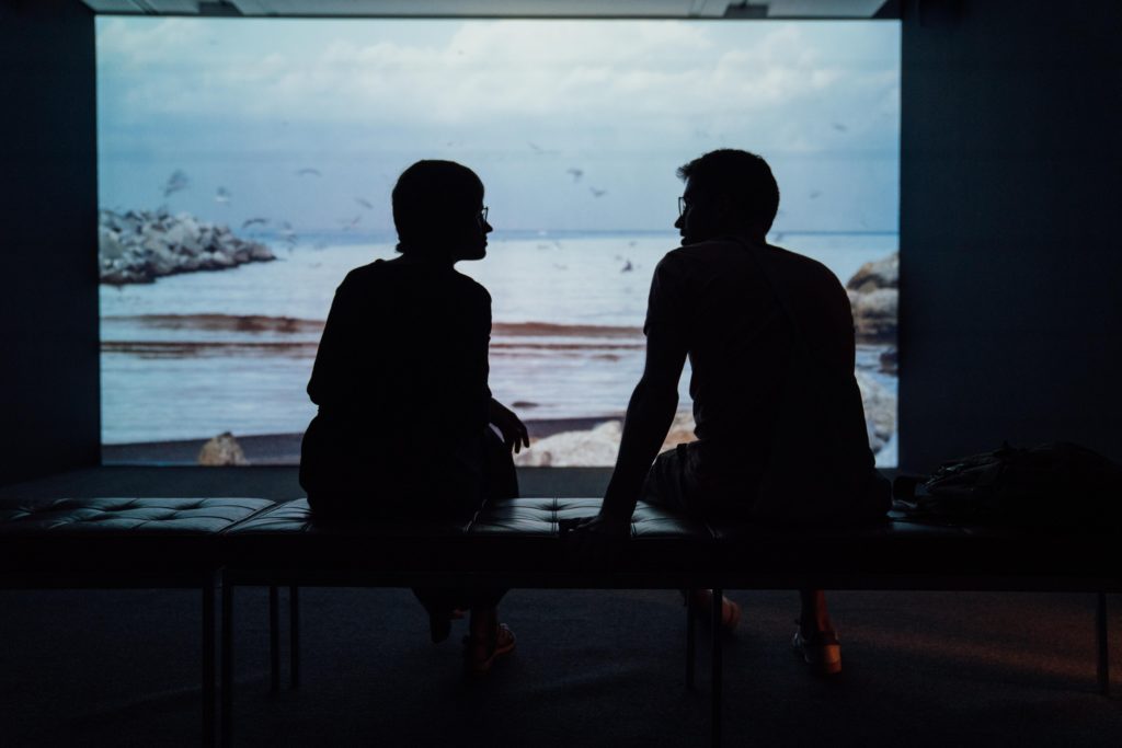silhouette of man and woman sitting on a bench with a screened image of a bay in the background