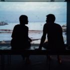 silhouette of man and woman sitting on a bench with a screened image of a bay in the background