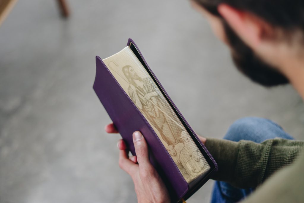 un hombre sostiene una Biblia encuadernada en cuero púrpura con la imagen de Jesús como humilde pastor en los bordes de la página