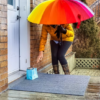 person with mustard colored jacket and rainbow umbrella leaves a blue gift bag on a neighbor's doorstep