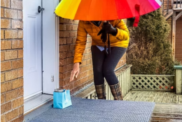 una persona con una chaqueta color mostaza y un paraguas arco iris deja una bolsa de regalo azul en la puerta de un vecino