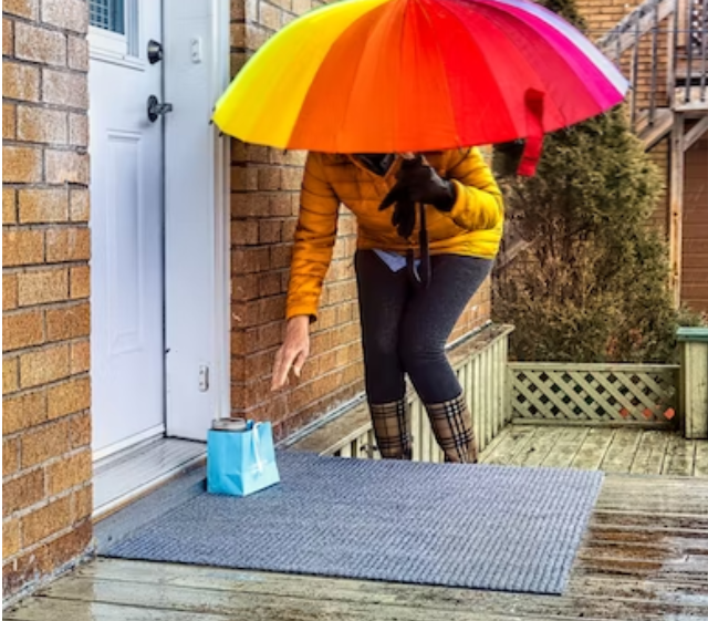 una persona con una chaqueta color mostaza y un paraguas arco iris deja una bolsa de regalo azul en la puerta de un vecino