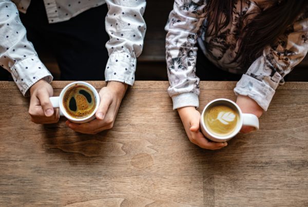 two people hold coffee cups over a wooden table, one with an impartial hand