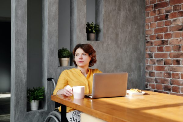 woman in a wheelchair looks off in the distance while sitting at a table with laptop
