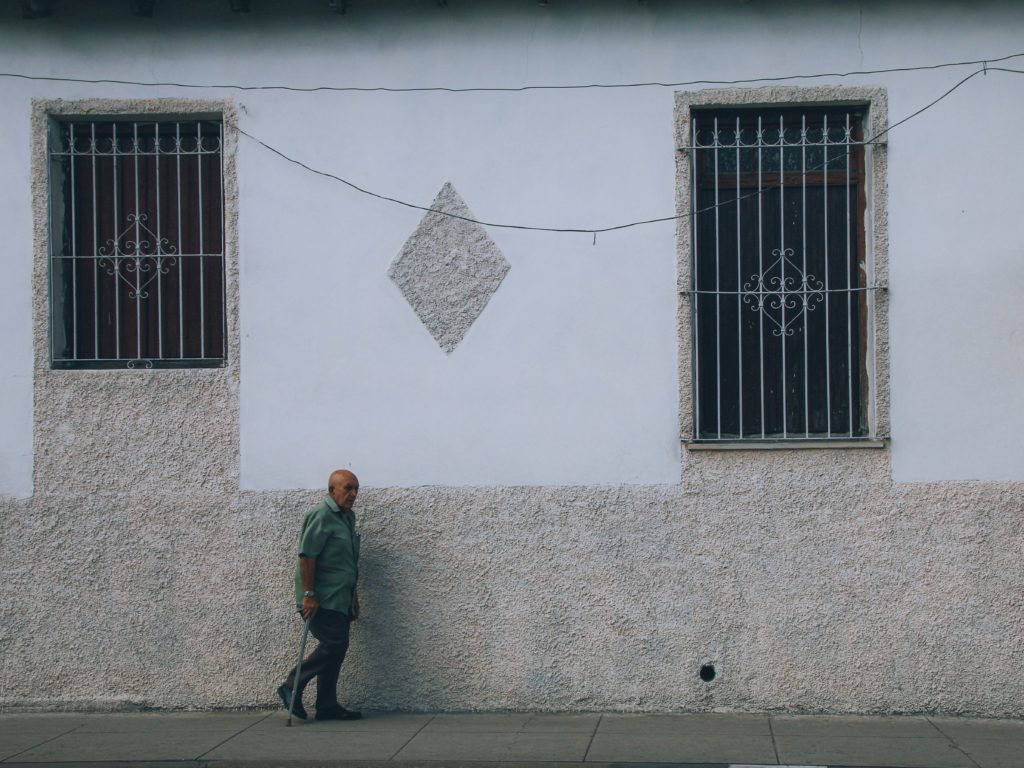man walks with a cane in front of a cement wall with barred windows