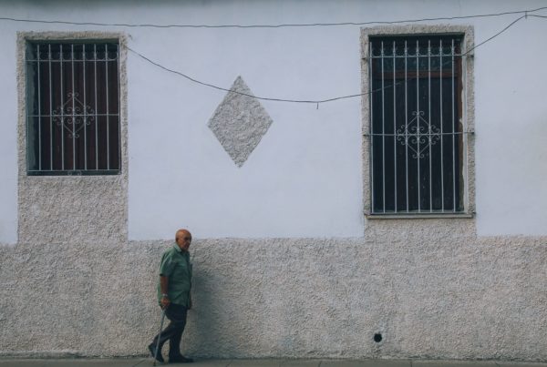 un hombre camina con un bastón frente a un muro de cemento con ventanas enrejadas