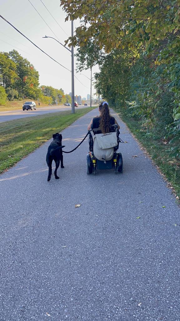 una mujer en silla de ruedas rueda por la acera con su perro negro al lado