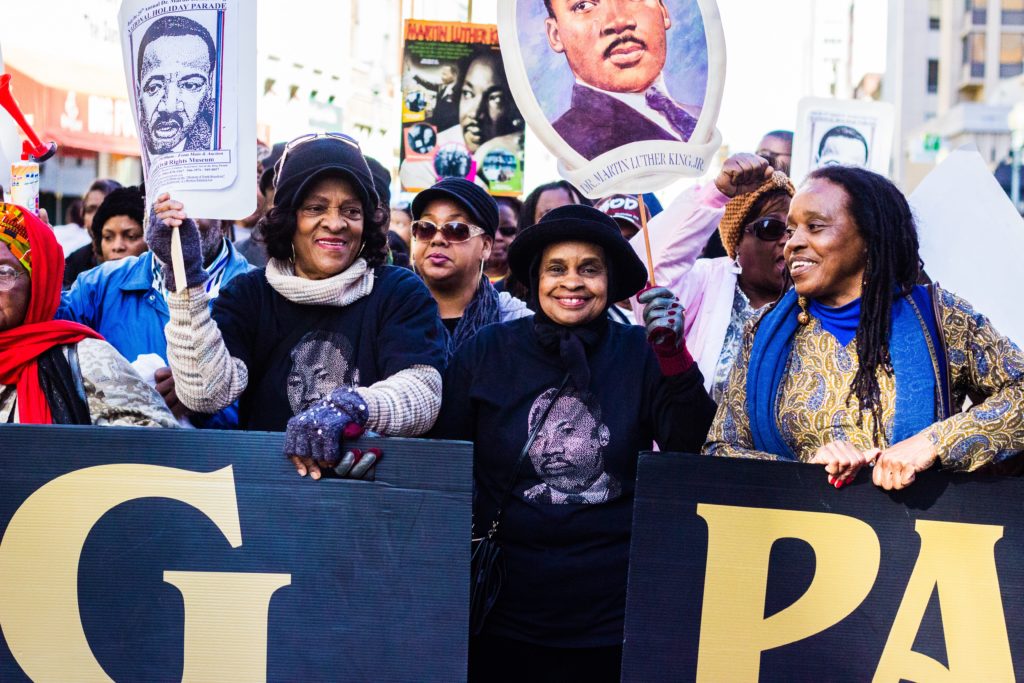 three Black women marchers smile and hold signs during MLK Day parade