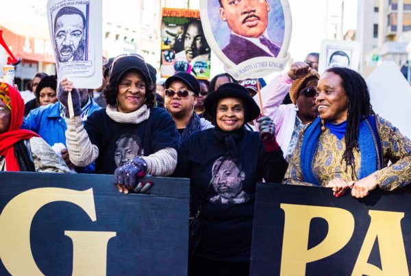 tres manifestantes negras sonríen y sostienen pancartas durante el desfile del Día MLK