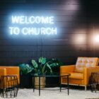two cozy chairs, table, and plant in front of neon "welcome to church" sign