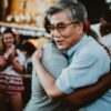 hombre asiático sonriente recibe el abrazo de una persona negra con gente aplaudiendo de fondo