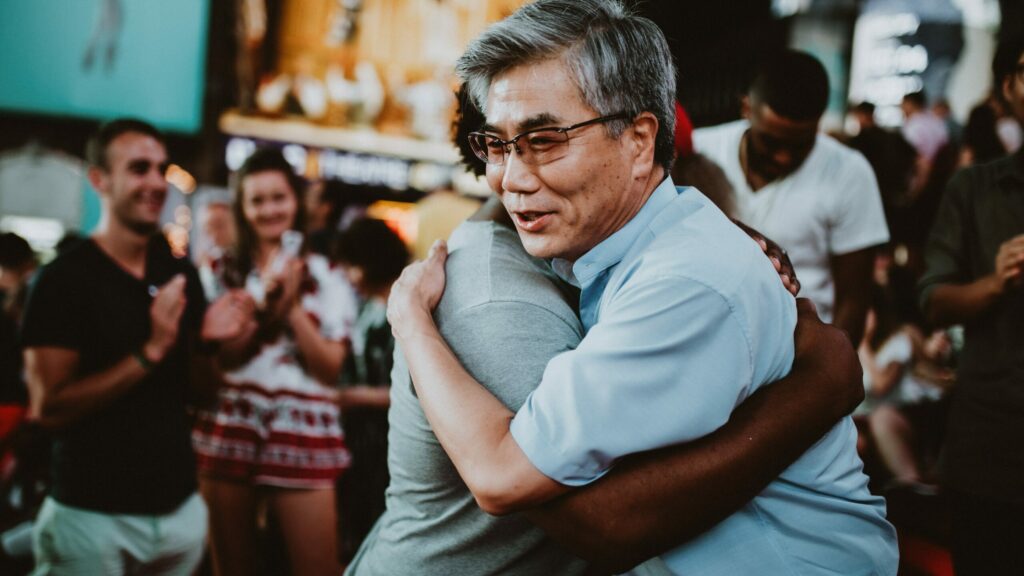 hombre asiático sonriente recibe el abrazo de una persona negra con gente aplaudiendo de fondo