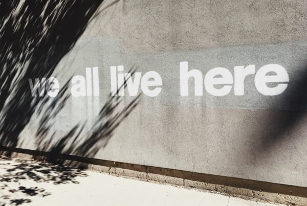gray cement wall with words "we all live here" spray painted in white
