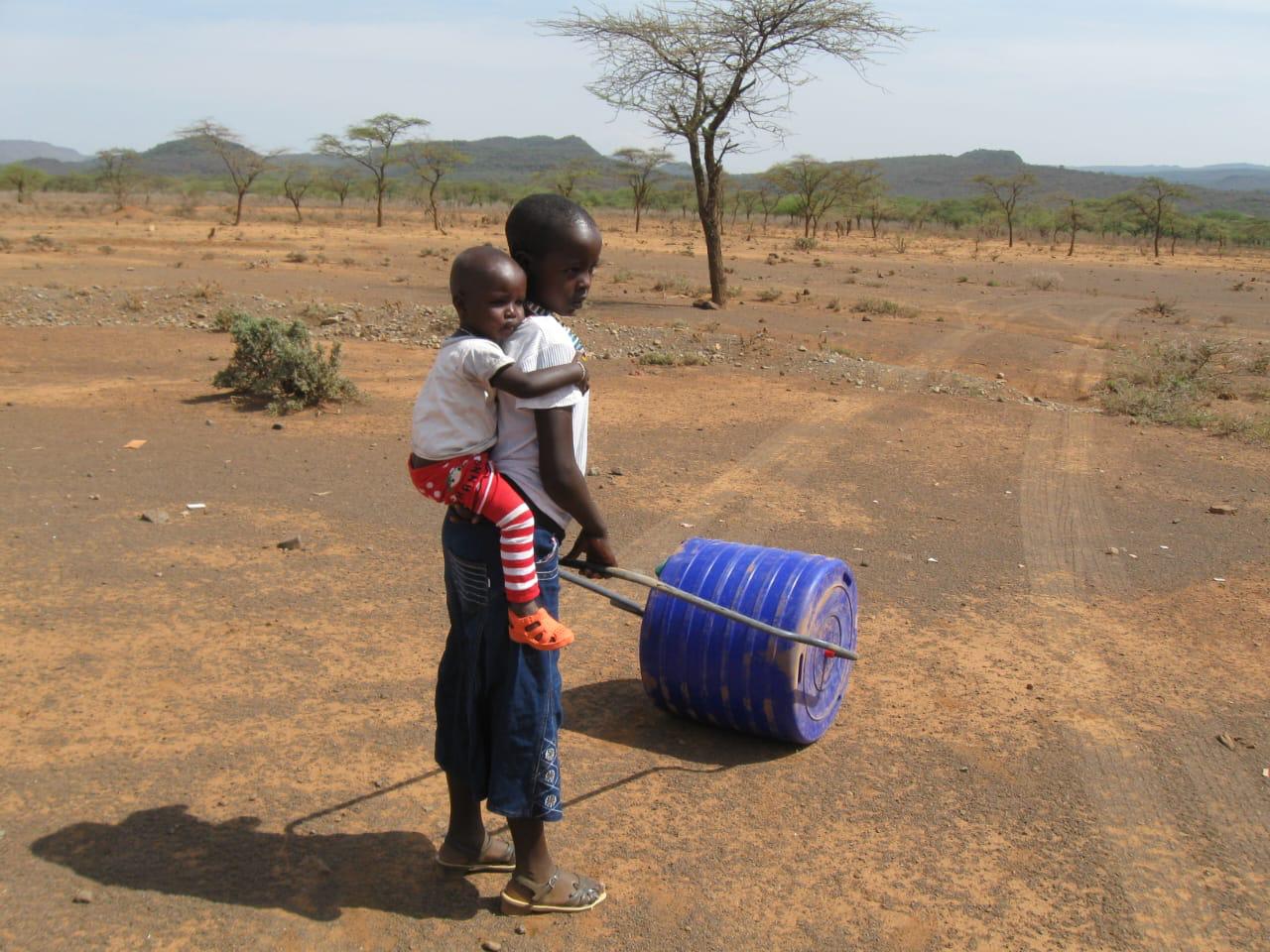 young African girl pushing blue water roller with younger brother on her back